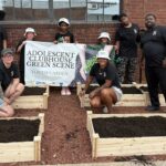 Member of the Adolescents Club House posed around newly erected raised garden beds with banner that says "Adolescents Clubhouse Green Scene"