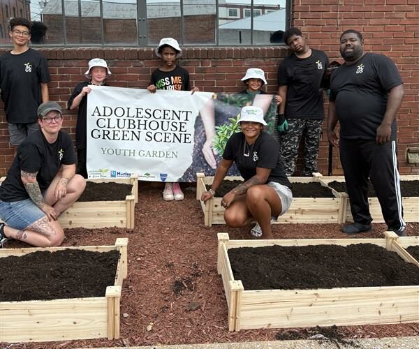 Member of the Adolescents Club House posed around newly erected raised garden beds with banner that says "Adolescents Clubhouse Green Scene"