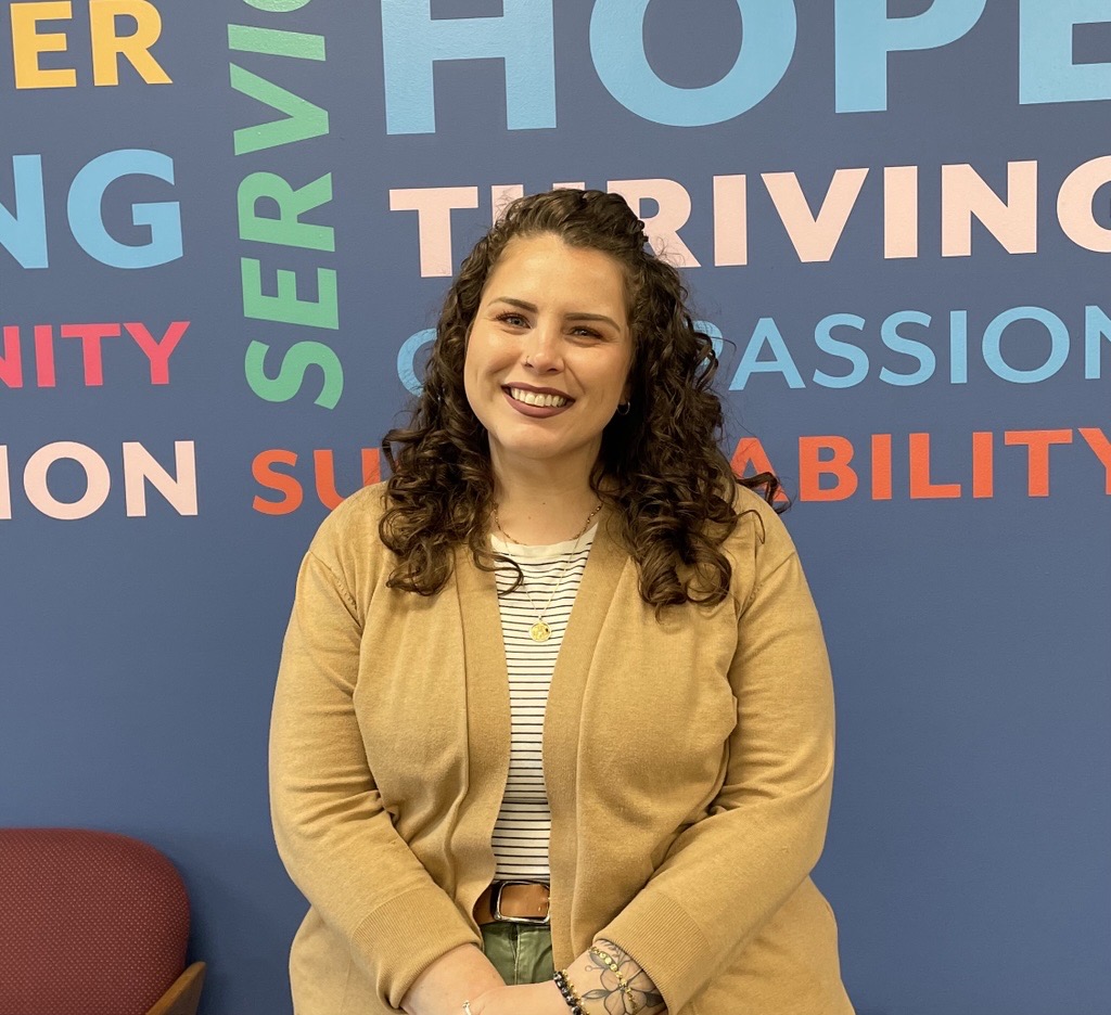 Allison Palmer, Chief Mission Officer of Horizon Goodwill, smiling with curly brown hair wearing a tan cardigan and striped shirt, standing in front of a blue wall with colorful words including "Hope," "Thriving," and "Compassion."